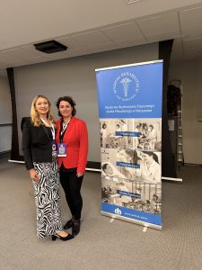 dr Dominika Zawadzka oraz mgr Anna Szweda, stoją uśmiechnięte na dla plakatu Wydziału Fizjoterapii AWF Warszawa. Ania ubrana jest w czarną marynarkę i spodnie w moty zebry, Dominika w czerwoną marynarkę i czarne spodnie